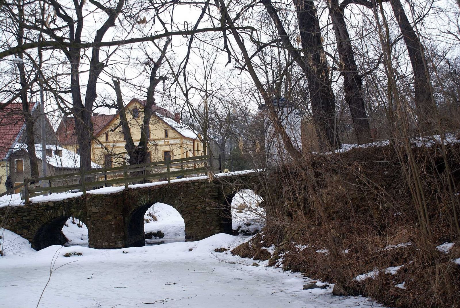 Kamienny most z trzema łukami nad zamarzniętą wodą w Bagieńcu, w tle zabudowania wsi.