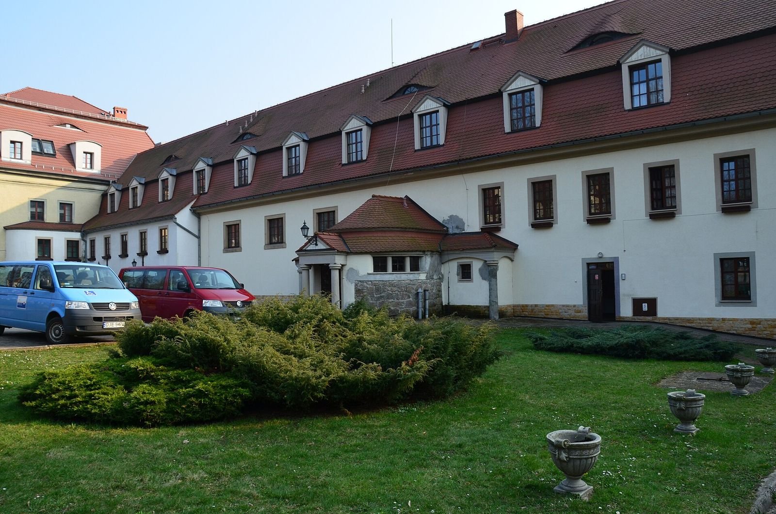 Biała elewacja pałacu w Janowicach Wielkich z dachem z lukarnami, gankiem wejściowym i trawnikiem z kamiennymi wazonami.