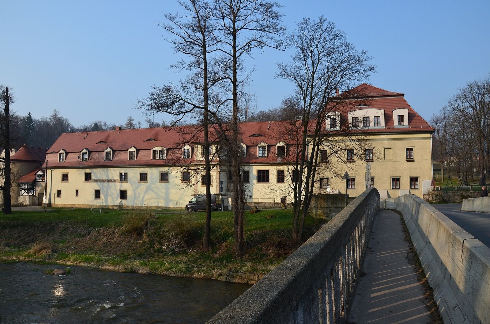 Pałac w Janowicach Wielkich widziany zza rzeki z mostku, z długim dachem z lukarnami i drzewami na pierwszym planie.