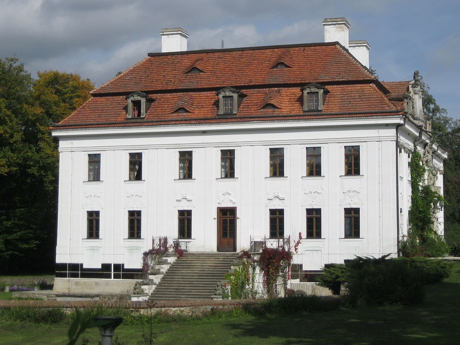 Dwukondygnacyjna jasna fasada pałacu z rzędami okien, schodami i czerwonym dachem mansardowym.