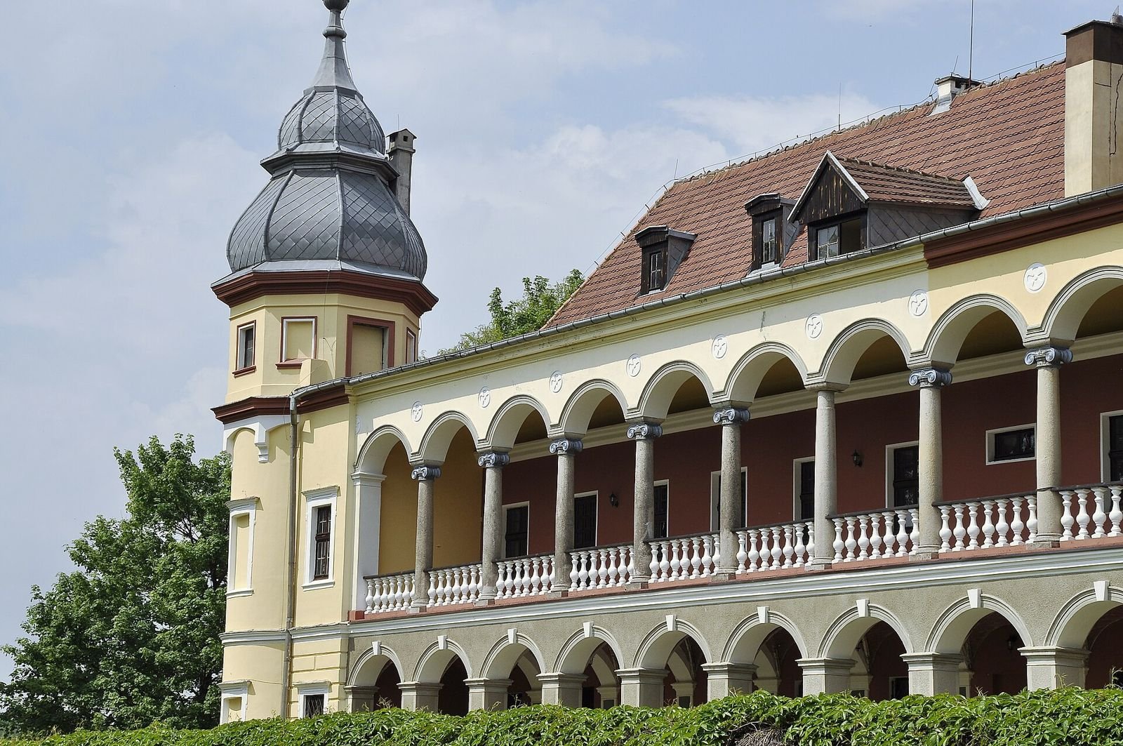 Dwukondygnacyjna weranda z arkadami i balustradą oraz narożna wieża z hełmem.