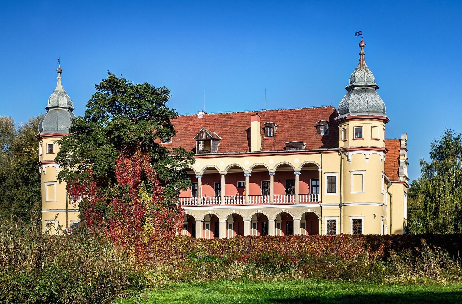 Żółty pałac z czerwonym dachem, dwiema narożnymi wieżami i dwukondygnacyjną loggią z arkadami, widziany od frontu spod błękitnego nieba.
