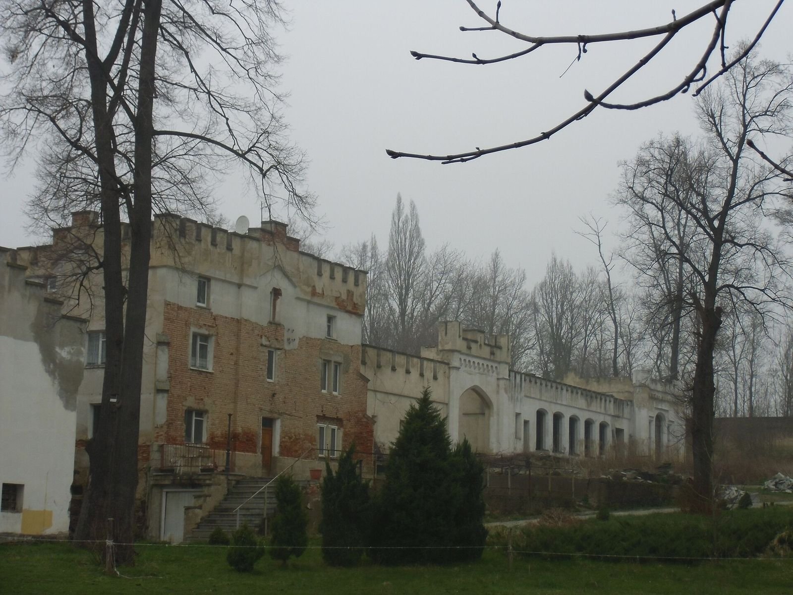 Ceglano-tynkowe zabudowania zespołu z blankami i arkadową galerią, wśród drzew parku.