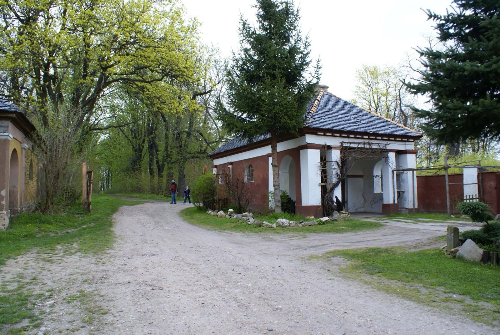 Murowany budynek bramny z kolumnami przy żwirowej drodze wjazdowej wśród drzew.