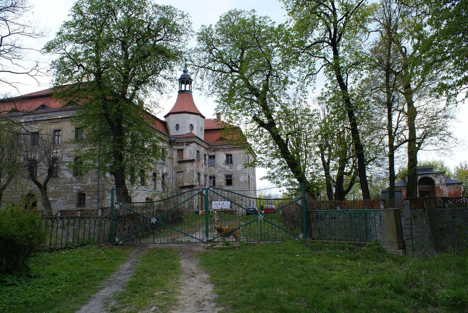 Pałac w Domanicach widoczny za zieloną metalową bramą i drzewami, od strony wjazdu.