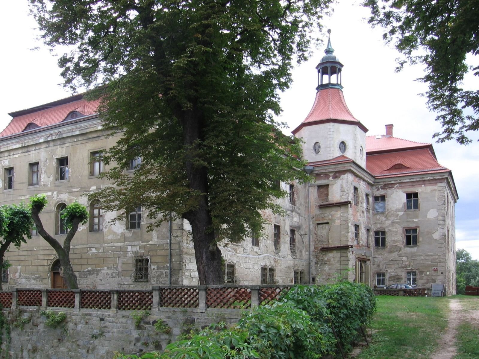Pałac w Domanicach z wieżyczką i czerwonymi dachami, widziany zza drzew i niskiego ogrodzenia.