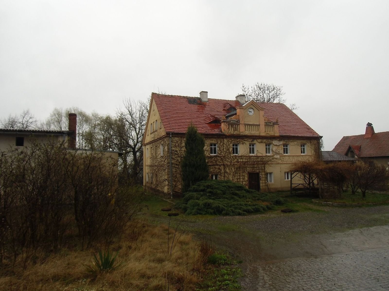 Dawny budynek gospodarczy zespołu pałacowego w Kłonicach z czerwonym dachem i zegarem w szczycie, przed nim drzewka iglaste i żwirowy dziedziniec.