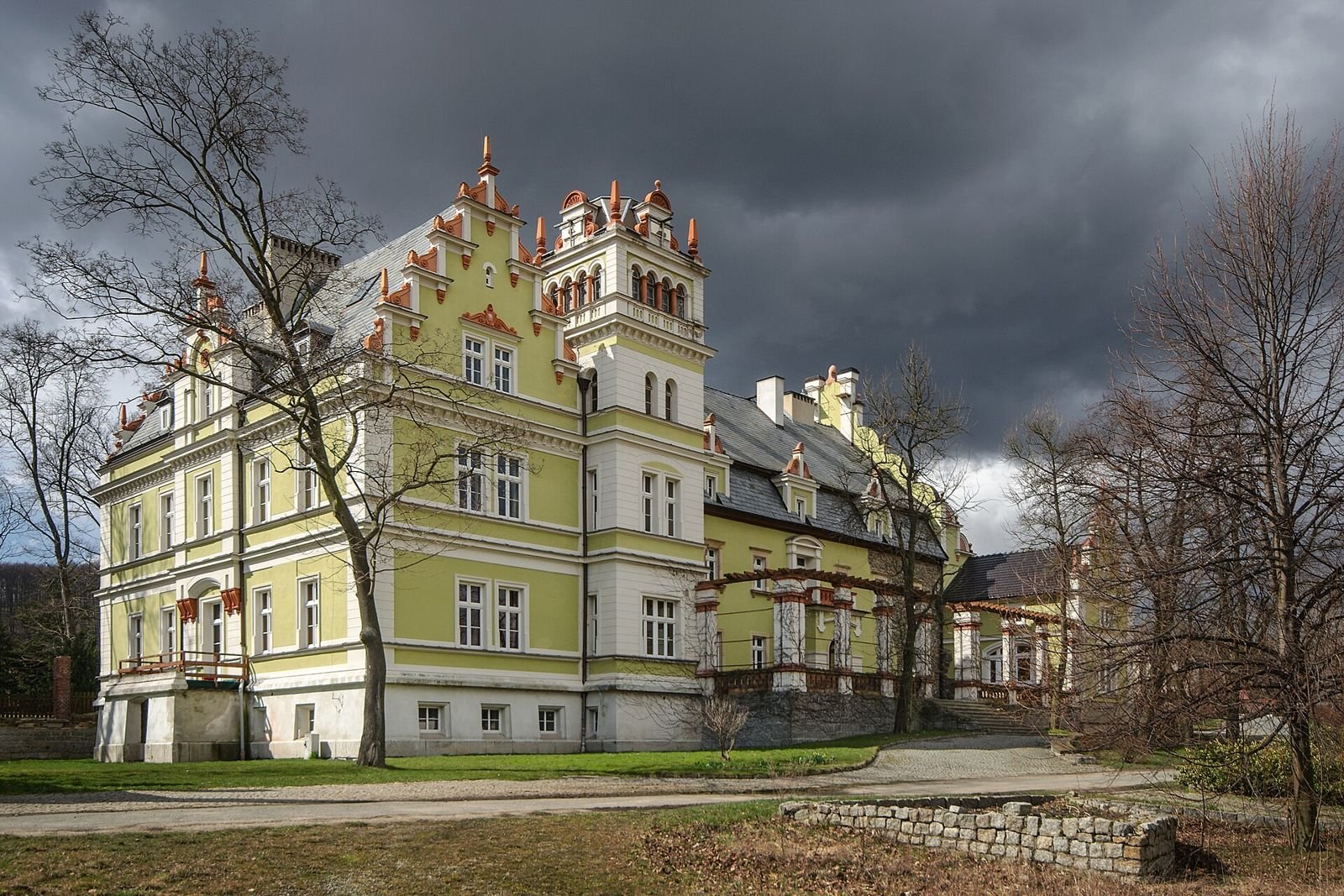 Pałac w Kłonicach widziany z narożnika, z wieżyczką, ozdobnymi szczytami i bocznym skrzydłem, na tle ciemnych chmur.