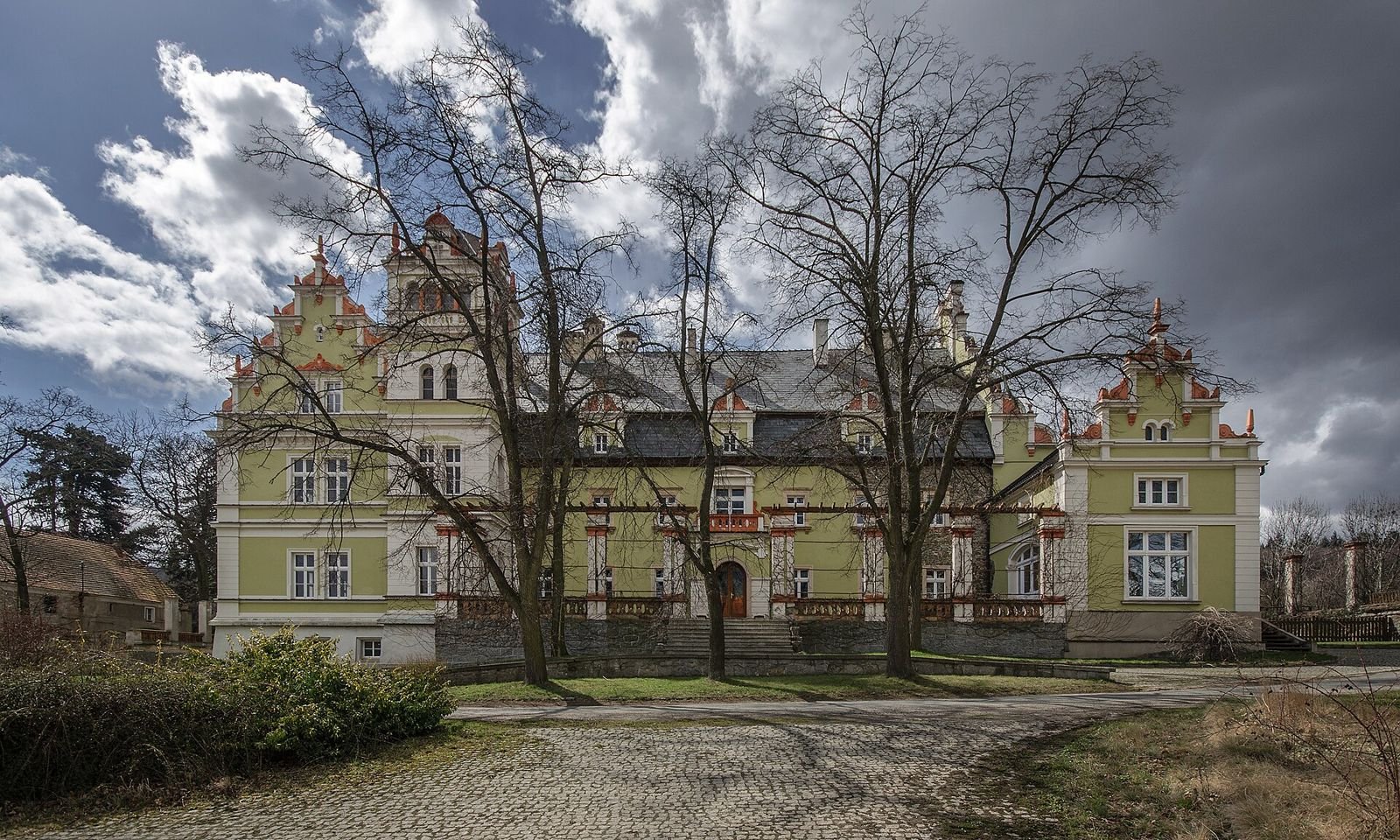 Pałac w Kłonicach widziany od frontu: jasnozielona elewacja z czerwonymi detalami, ozdobne szczyty i wieża, na pierwszym planie bruk i bezlistne drzewa.