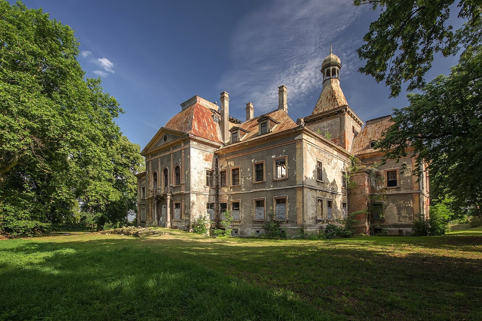 Pałac w Mańczycach widziany z ukosa od strony trawnika, z wieżą, wysokimi dachami i kominami.