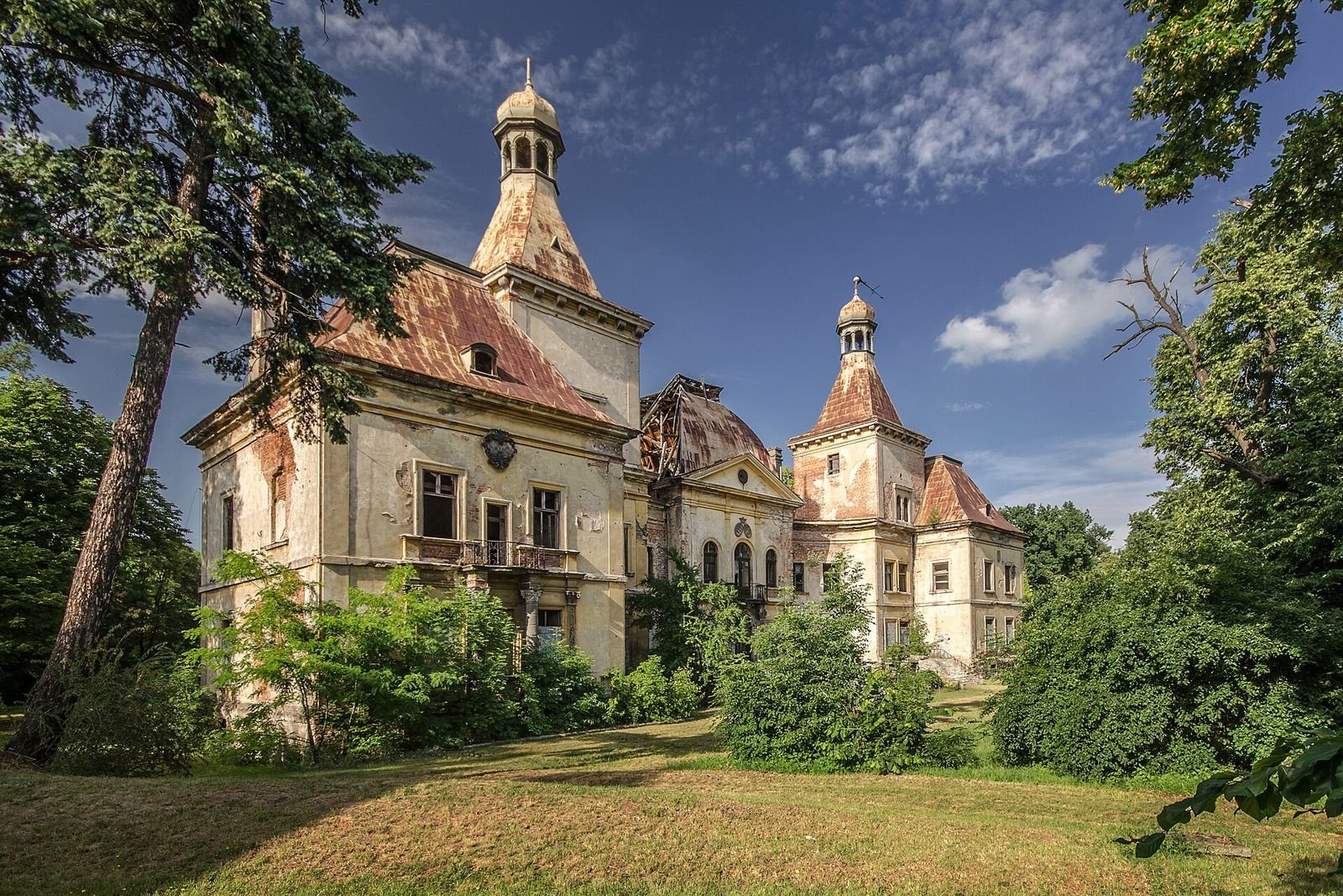 Pałac w Mańczycach widziany z boku spomiędzy drzew, z balkonem, wieżami i zniszczonymi dachami.