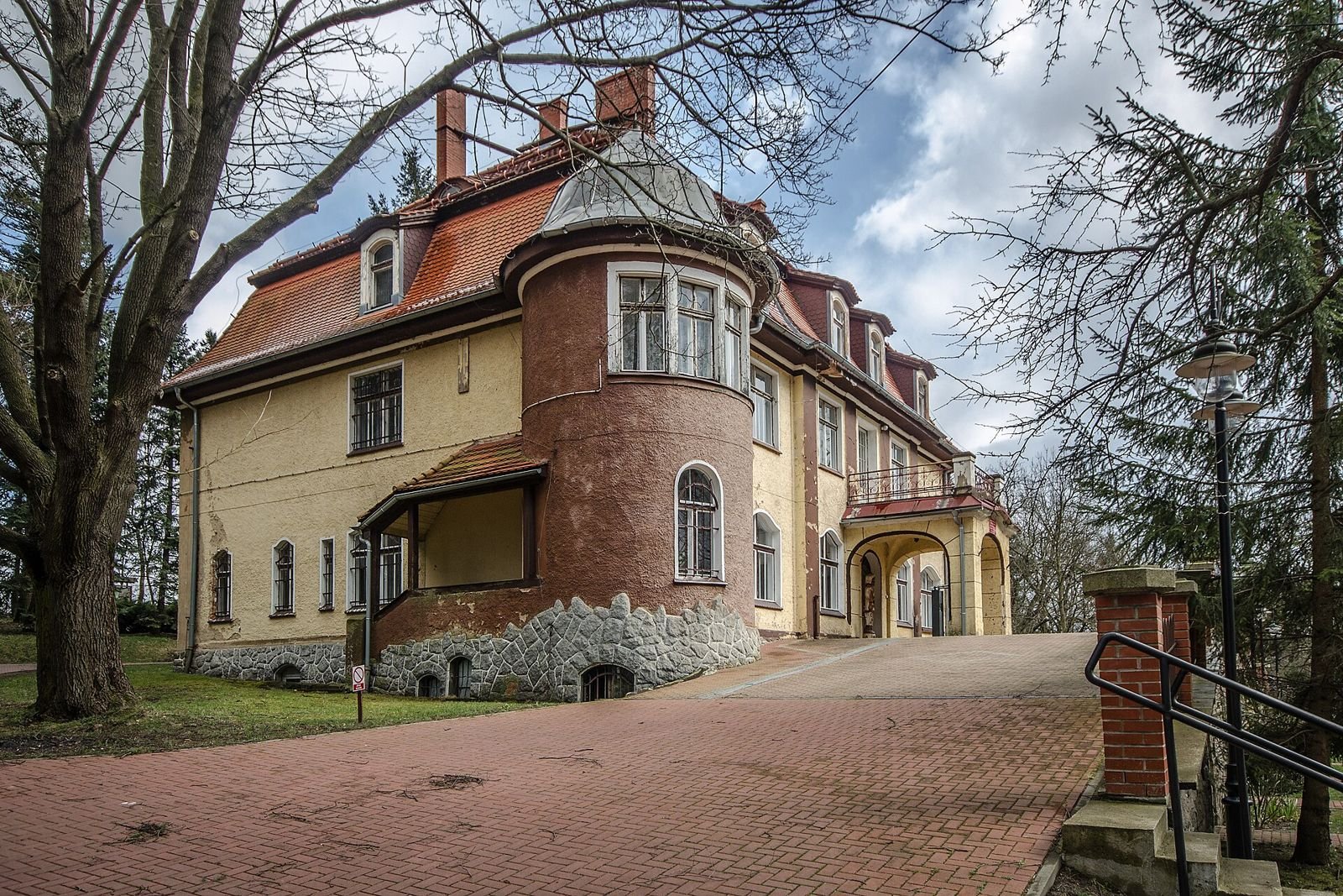 Pałac w Muchowie od strony podjazdu: okrągły ceglany ryzalit, wejście w podcieniu z tarasem i wybrukowany dziedziniec.