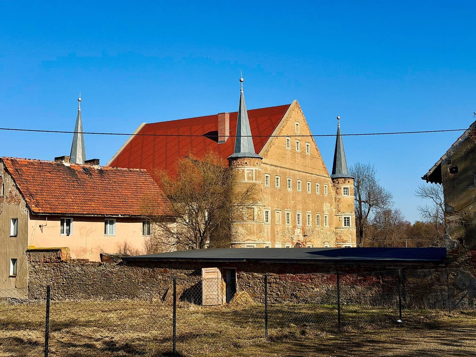 Dwór z basztami za zabudowaniami gospodarczymi folwarku.