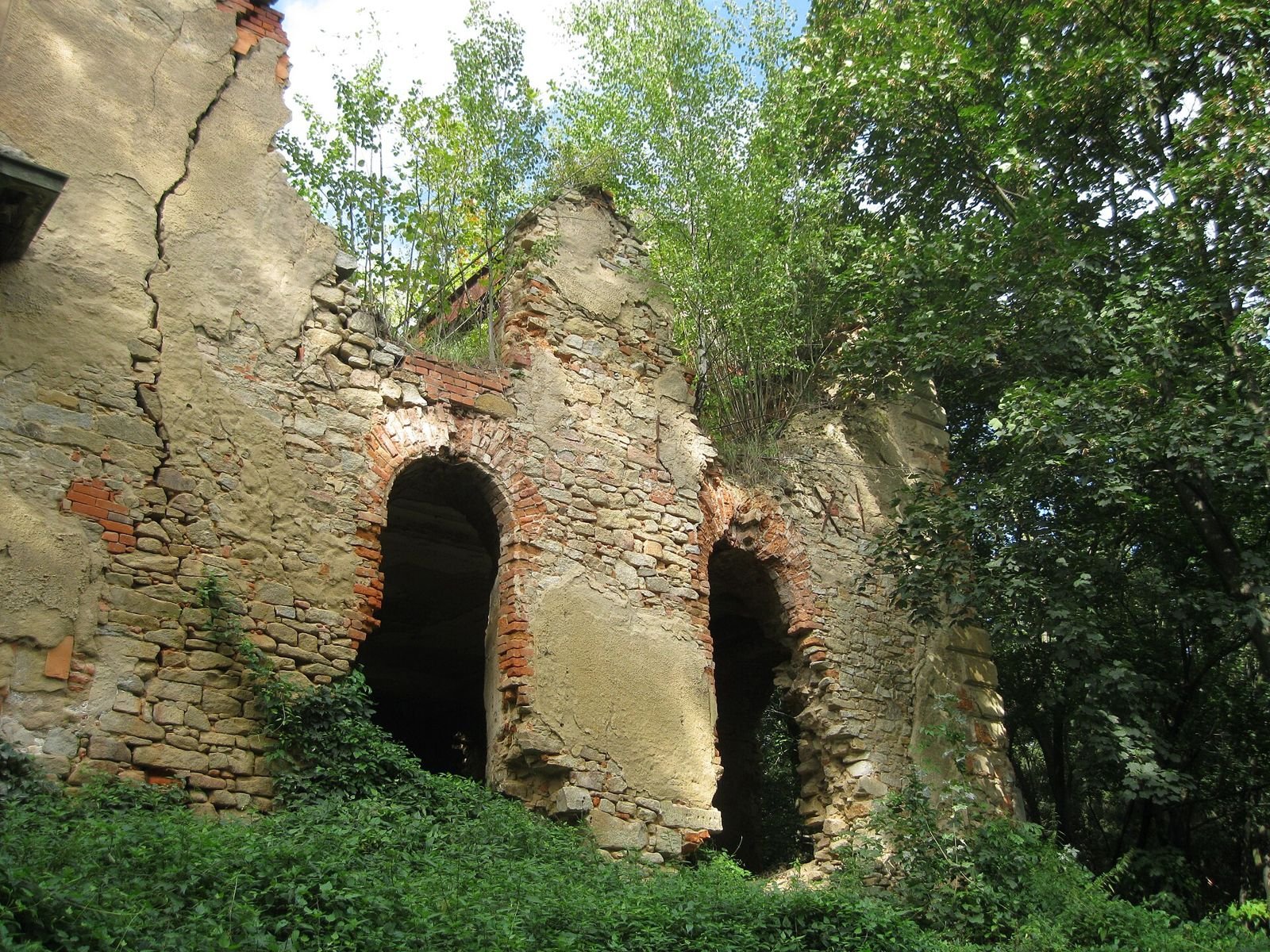 Fragment ruiny z kamiennego i ceglanego muru z dwoma łukowymi otworami, otoczony roślinnością.