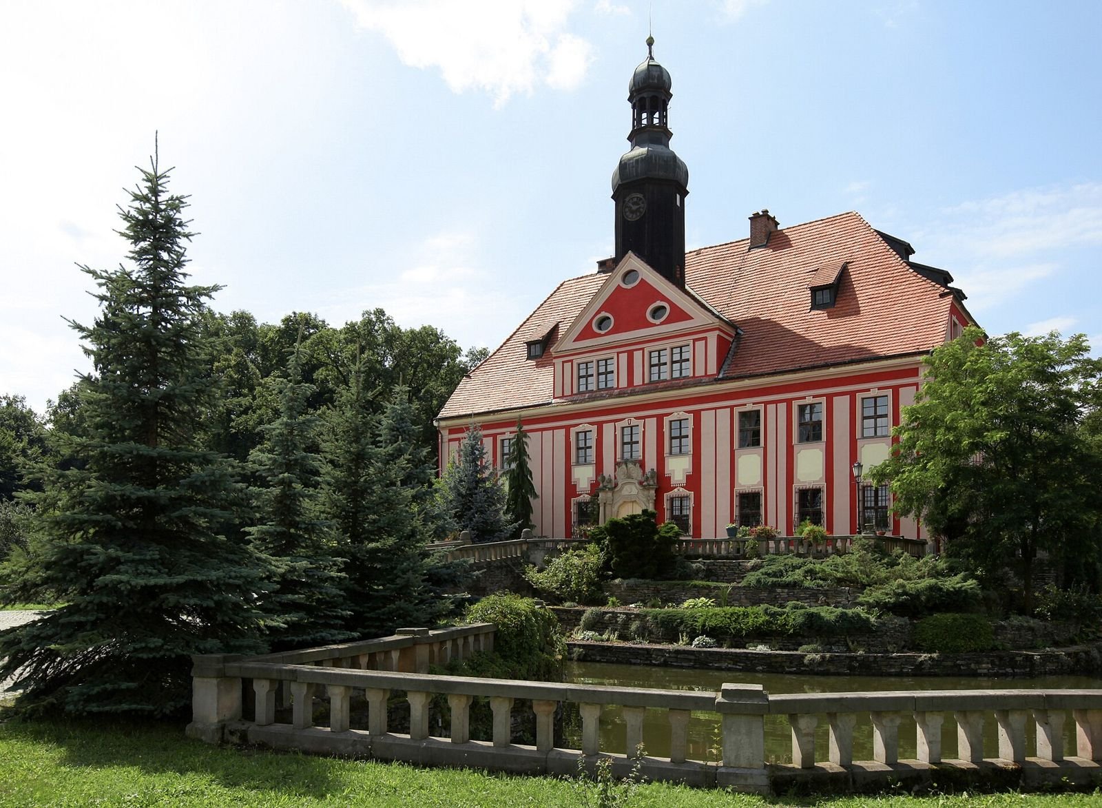 Barokowy pałac z czerwono-białą elewacją, wysokim dachem i wieżyczką, widziany zza kamiennej balustrady i drzew.