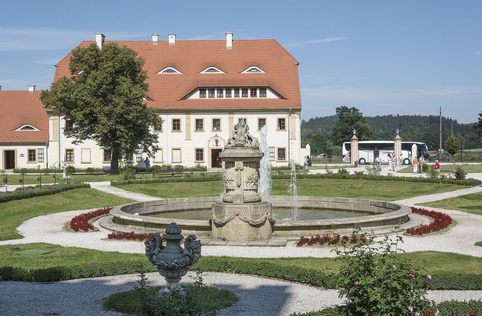Kamienna fontanna w okrągłej sadzawce, za nią budynek z czerwonym dachem i ogród z rabatami.