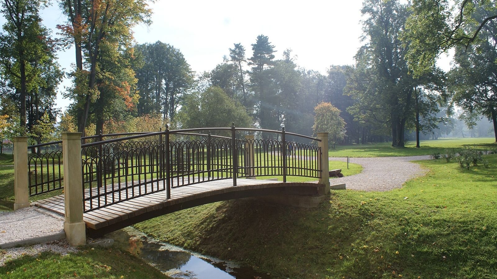 Metalowa kładka nad strumieniem w parku, wśród wysokich drzew i trawników.