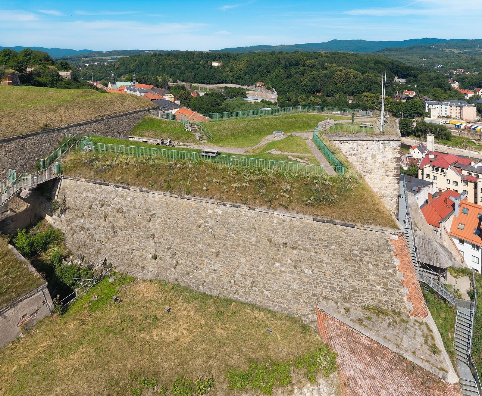 Bastion z tarasem widokowym Twierdzy Kłodzko z lotu ptaka, za nim dachy Kłodzka i zalesione wzgórza.
