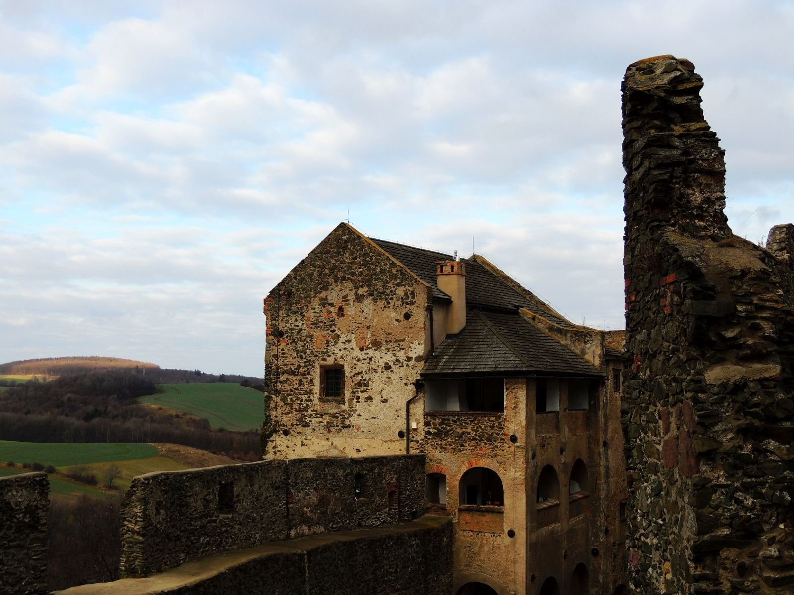 Kamienny budynek zamku w Bolkowie z arkadowym gankiem, obok mur i fragment ruin, w tle wzgórza