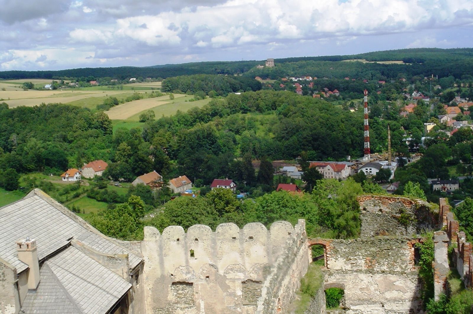 Widok z wieży zamku w Bolkowie na dachy i blanki zamku oraz pola i zalesione wzgórza w oddali.