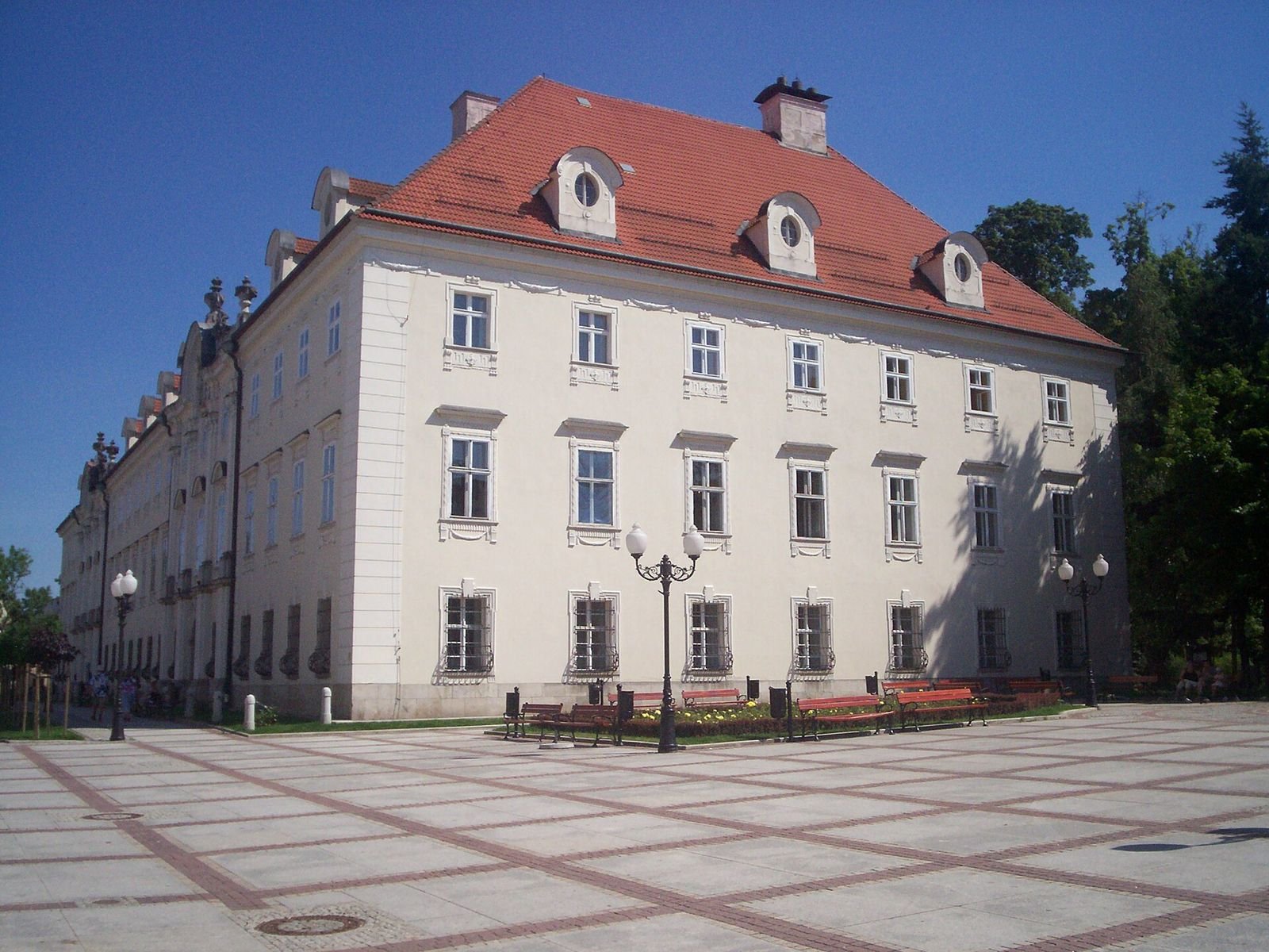 Narożnik pałacu z wysokim dachem czerwoną dachówką i lukarnami, przed nim wybrukowany plac z ławkami.