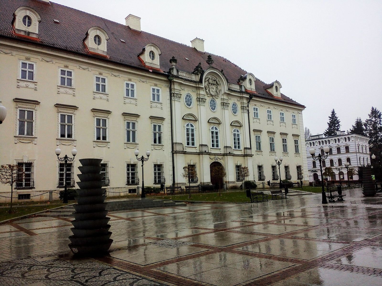 Wybrukowany plac przed pałacem z latarniami i ławkami, w tle fasada z portalem środkowym.