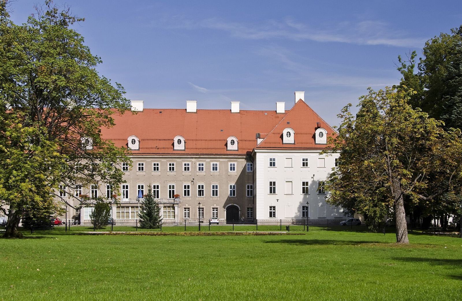 Pałac od strony parku: długa bryła z czerwonym dachem i lukarnami, przed nią trawnik i drzewa.