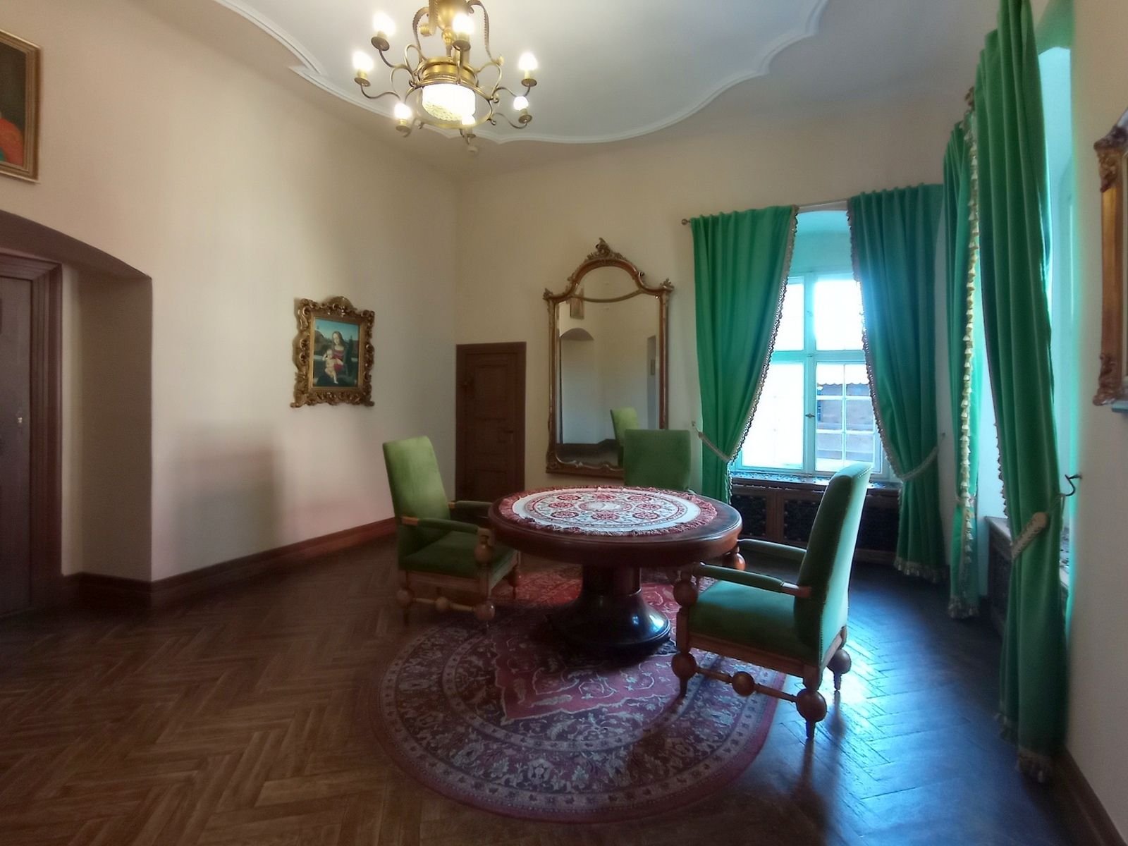 Komnata w Zamku Czocha z okrągłym stołem, zielonymi fotelami, zielonymi zasłonami i lustrem w złoconej ramie.