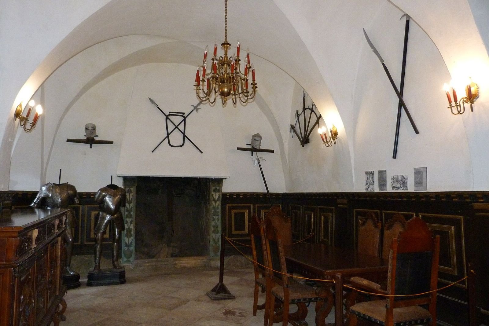 Sklepiona sala zamku Grodno ze zbrojami, bronią na ścianach, żyrandolem i stołem z krzesłami.