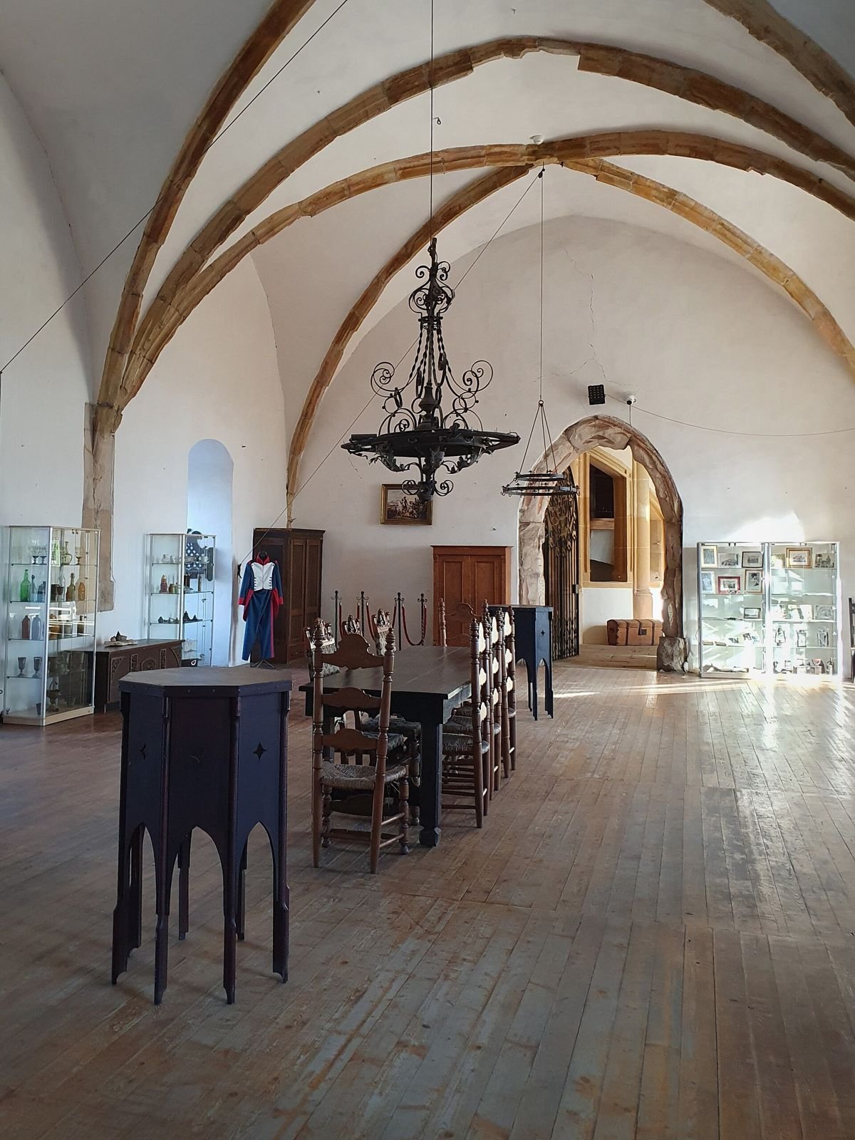 Wysoka sala zamku Grodziec z łukowym sklepieniem, kutym żyrandolem, długim stołem i gablotami pod ścianami.