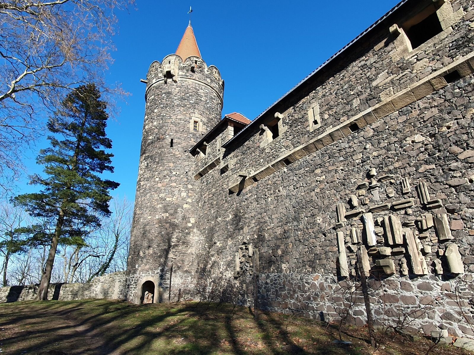 Mur zamku Grodziec z wmurowanymi kamiennymi detalami lapidarium, w głębi okrągła baszta z czerwonym stożkowym dachem.