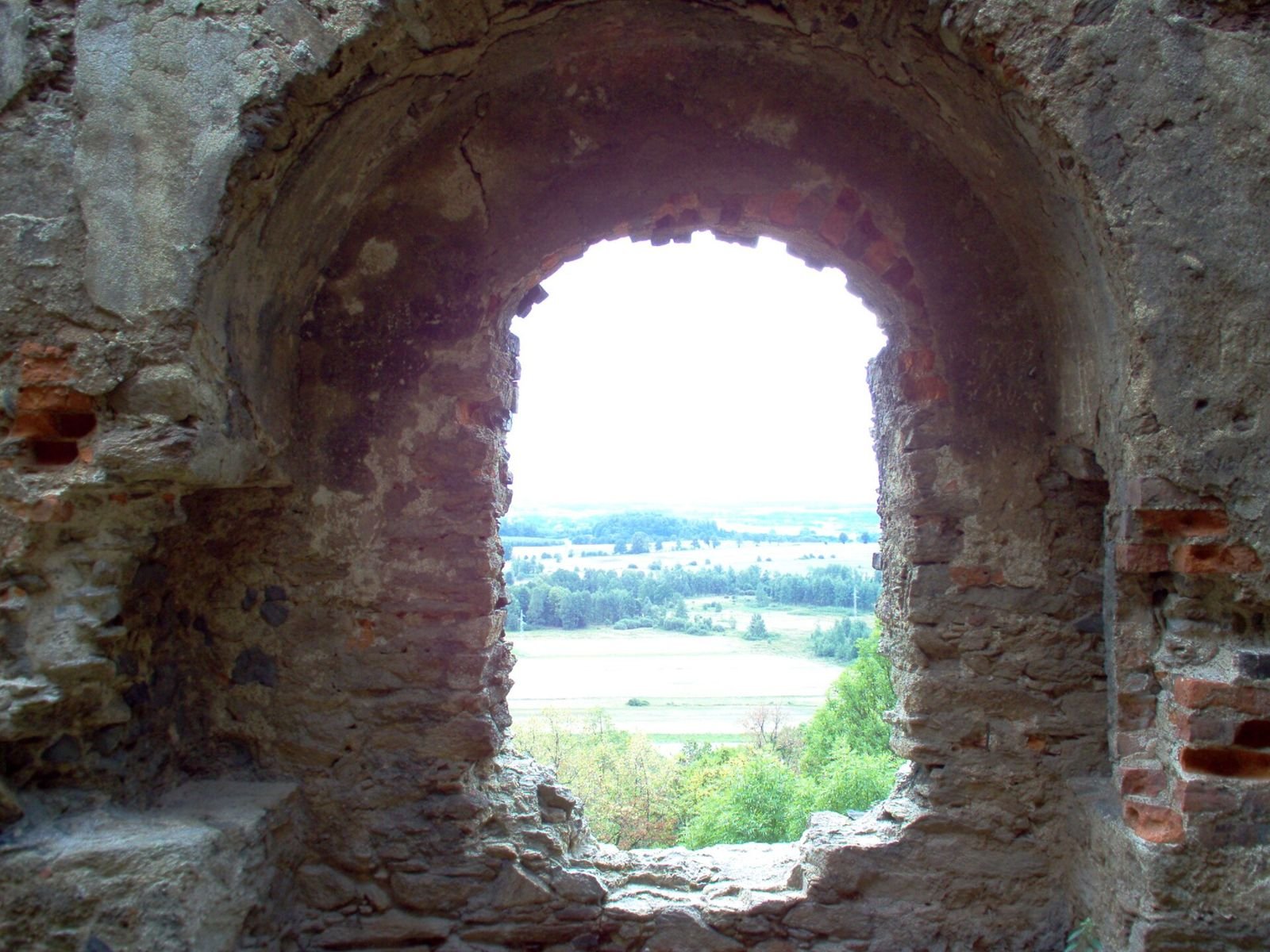 Widok przez sklepione okno ruin na pola i lasy w dolinie.