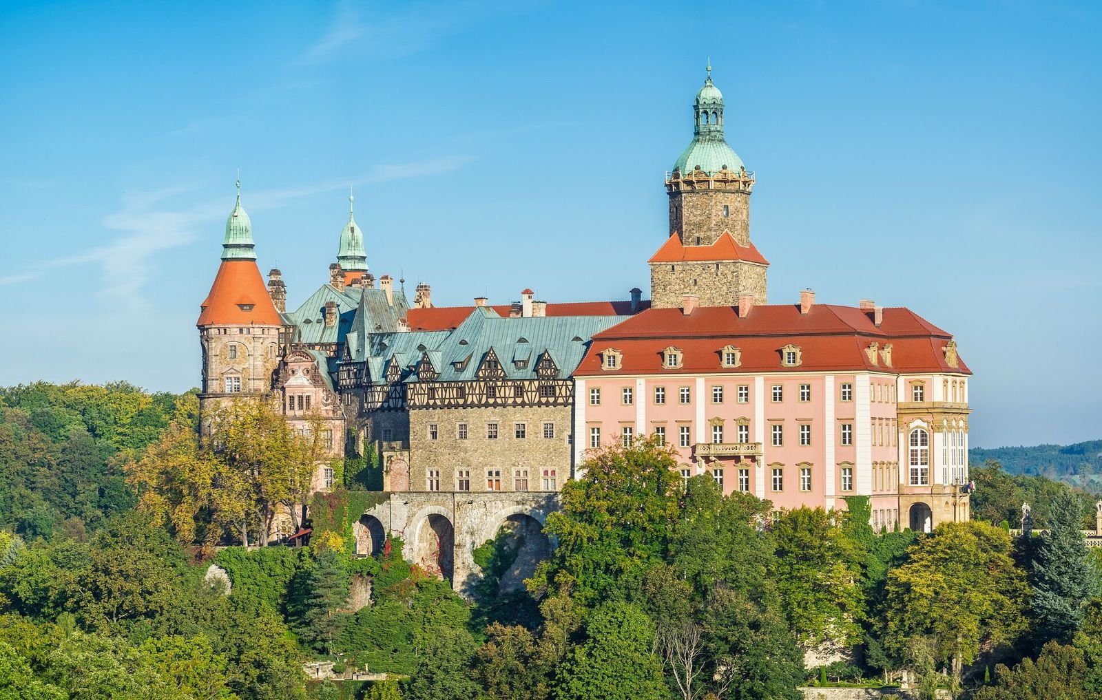 Zamek Książ w całości: różowe skrzydło, wieża z zielonym hełmem i kamienne mury ponad koronami drzew.