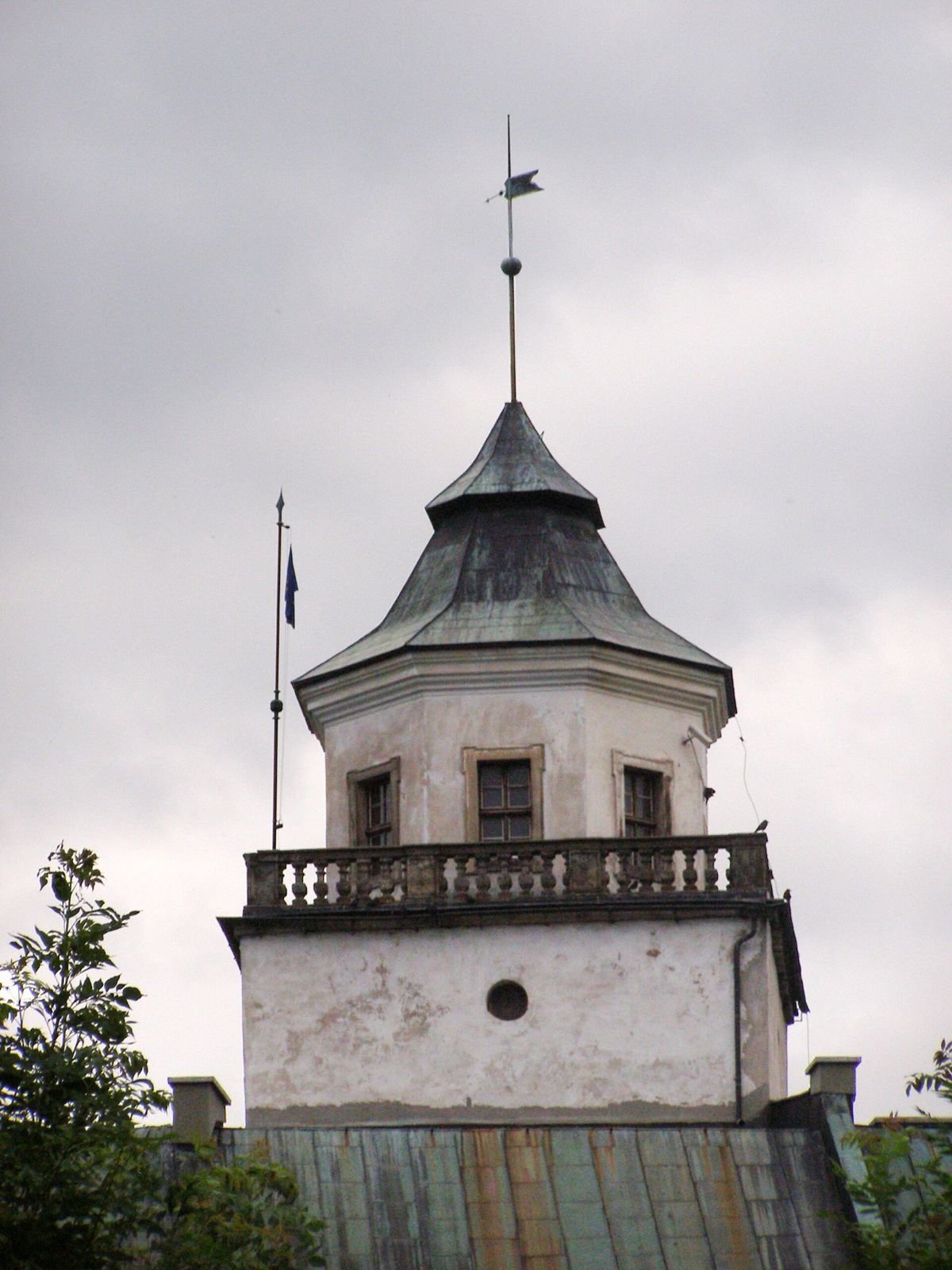 Górna część wieży z balkonem z balustradą, zielonym stożkowym dachem i chorągiewką na tle pochmurnego nieba.