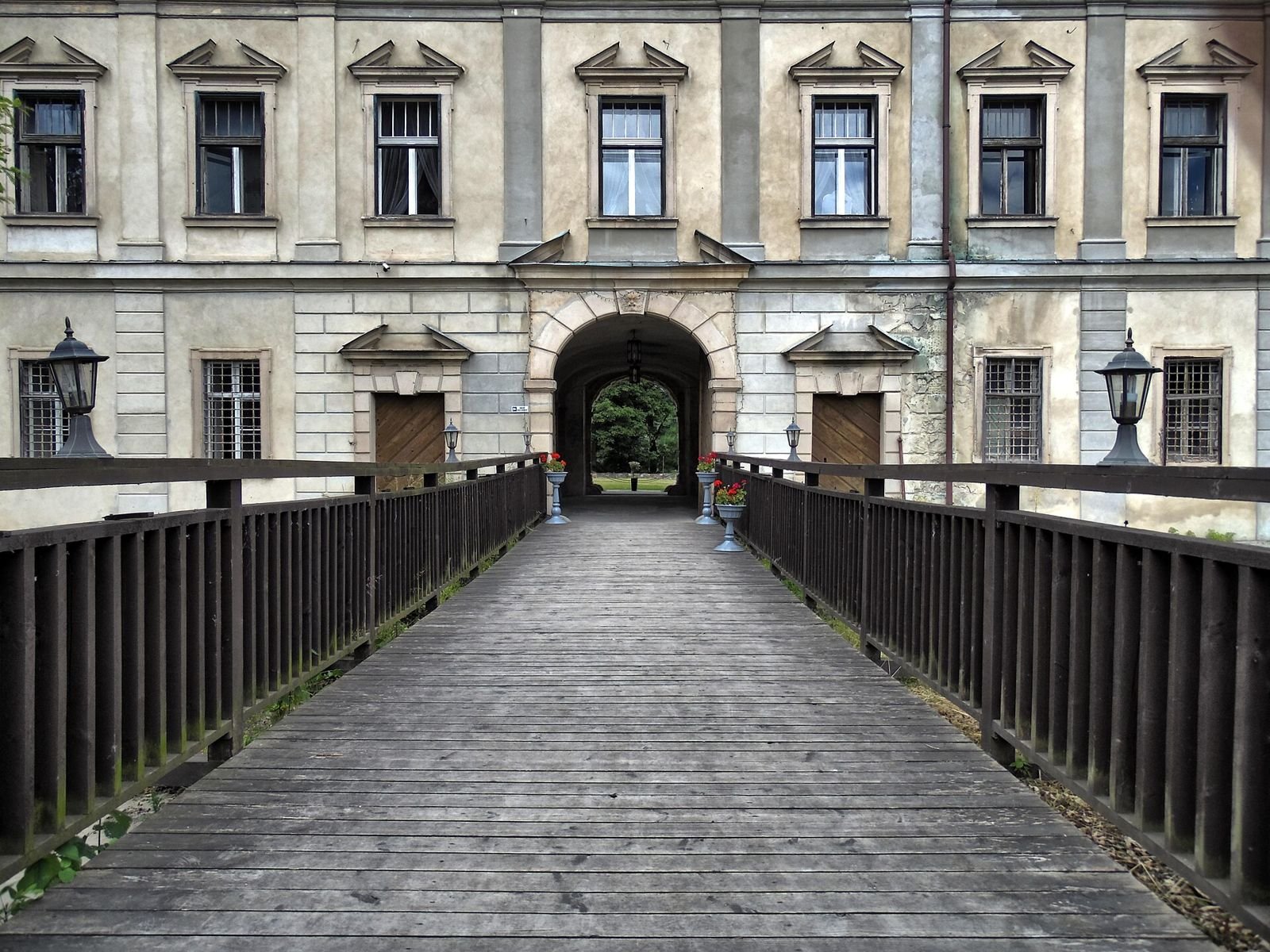Drewniany most prowadzący do bramy w elewacji zamku, po bokach balustrady i latarnie.