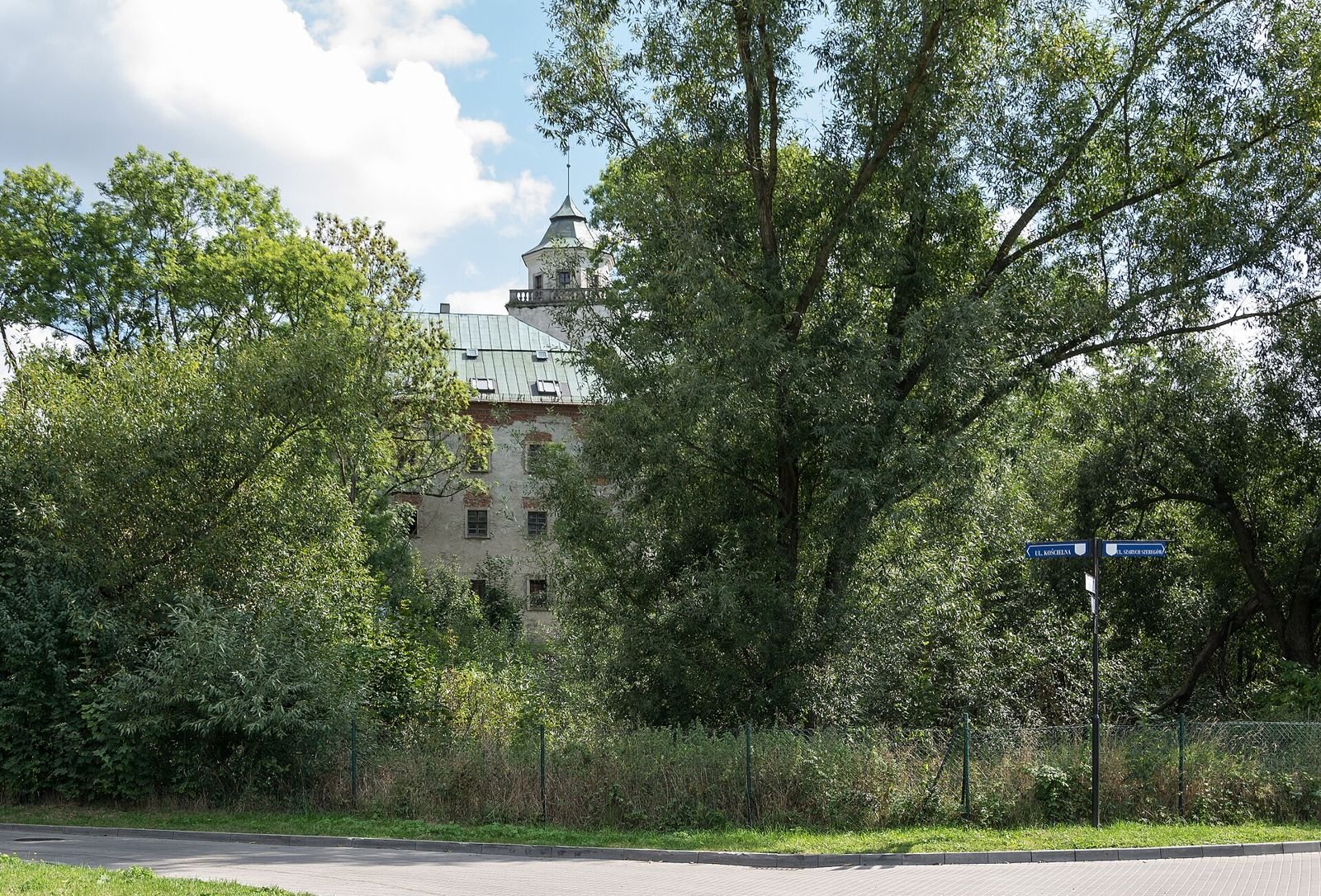 Zamek widoczny przez zieleń drzew i krzewów, na pierwszym planie droga i tablica drogowa.