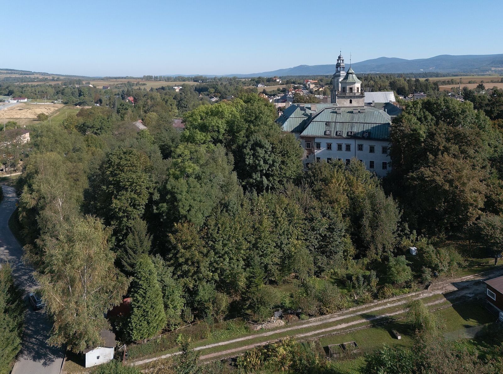 Zamek z wieżyczką wyłaniający się zza drzew na tle doliny i dalekich wzgórz, widok z dystansu z góry.