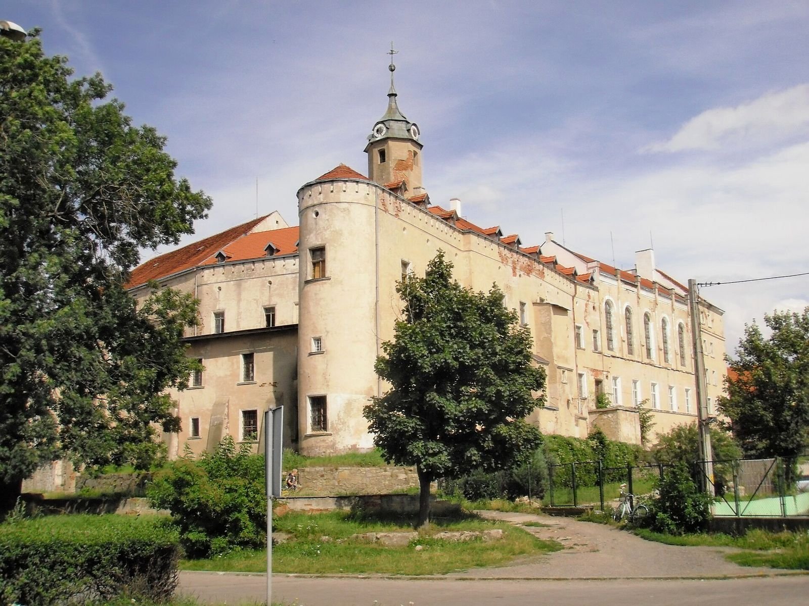 Zamek w Jaworze widziany z zewnątrz, z okrągłą basztą narożną i wieżyczką z zegarem nad dachami, wśród drzew.