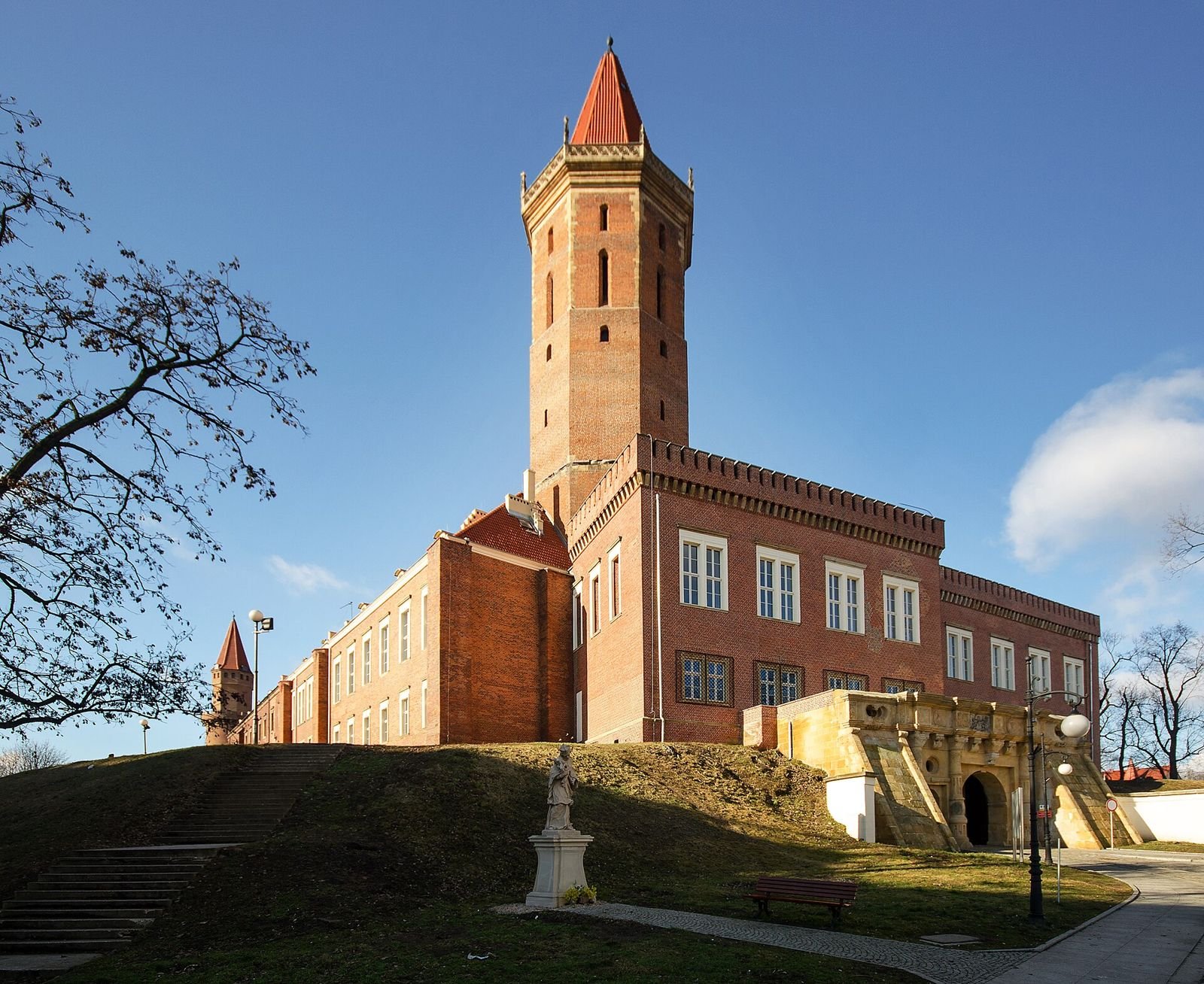 Ceglany zamek z wysoką okrągłą wieżą ze spiczastym hełmem, widziany od strony wzgórza w słoneczny dzień.