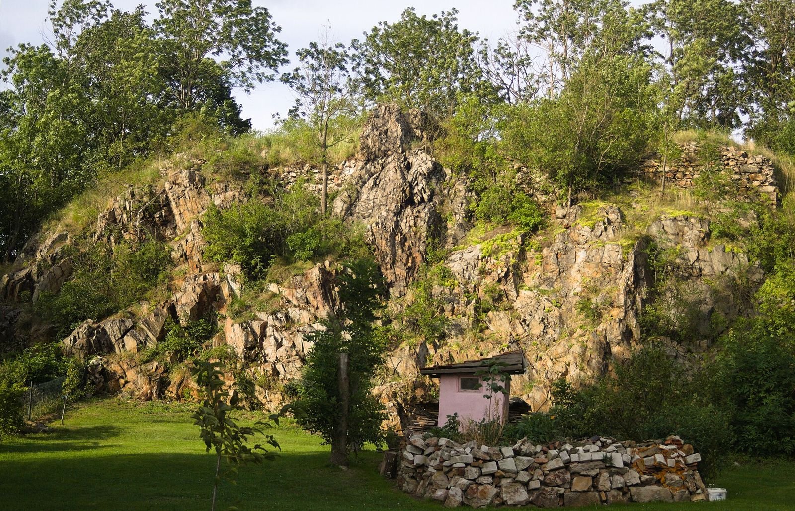 Skalna ściana z fragmentem ruin porośnięta zielenią, u jej podnóża mały domek i stos kamieni.