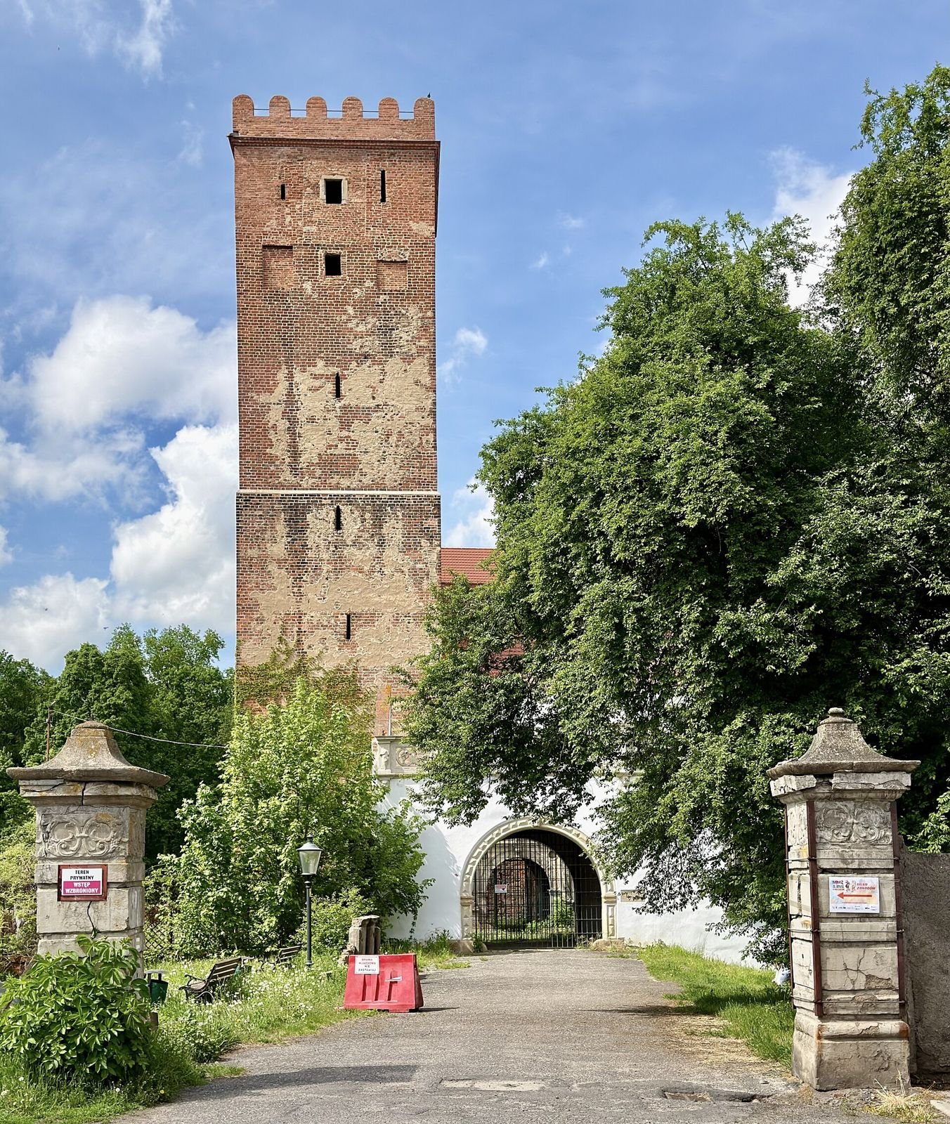 Ceglana wieża zamku w Prochowicach z krenelażem, widoczna za bramą wjazdową i kamiennymi słupami.