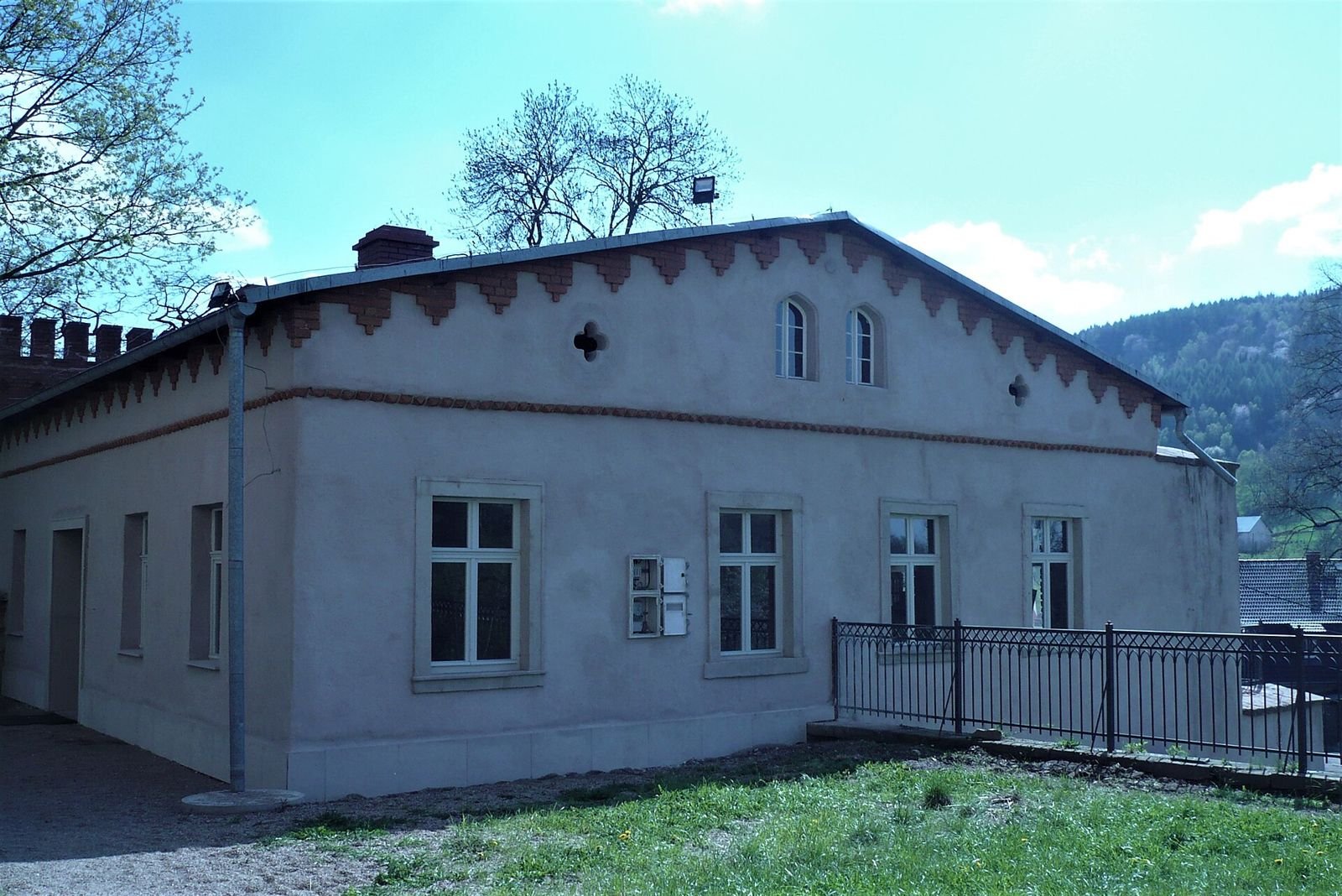 Parterowy budynek bramny z ozdobnym gzymsem i oknami, obok metalowa balustrada i trawnik.