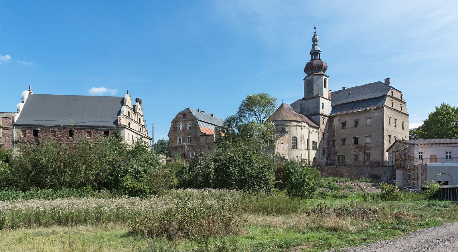 Widok z łąki na zespół pałacowy: wieża z baniastym hełmem, dawny spichlerz po lewej i pałac po prawej.