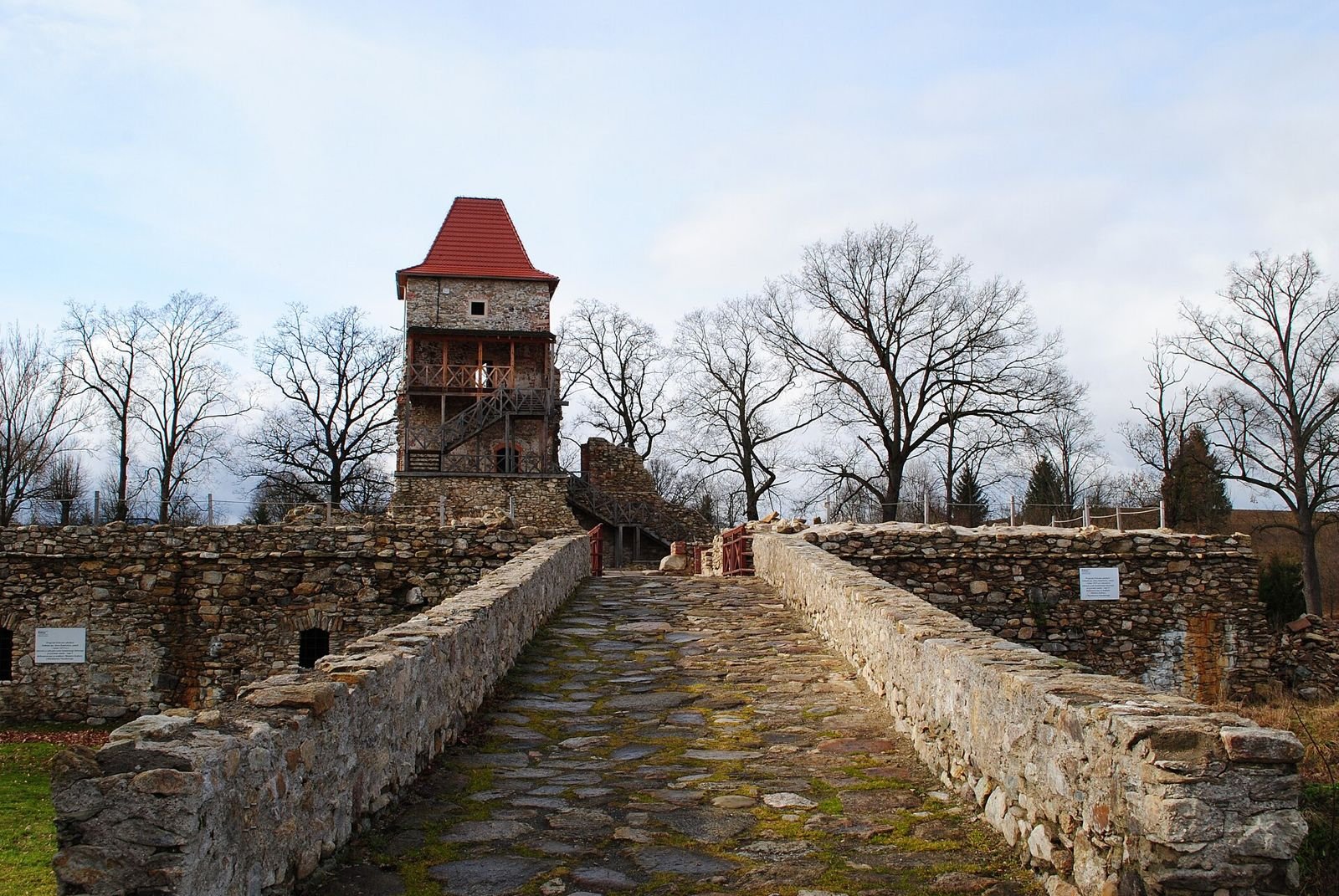 Brukowane przejście między niskimi kamiennymi murkami prowadzące do zamkowych murów i ceglasto-kamiennej wieży z czerwonym dachem i drewnianymi galeriami.