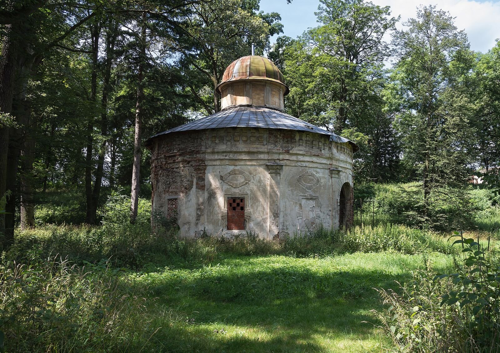 Okrągły pawilon parkowy z kopułką, otoczony drzewami i trawą.