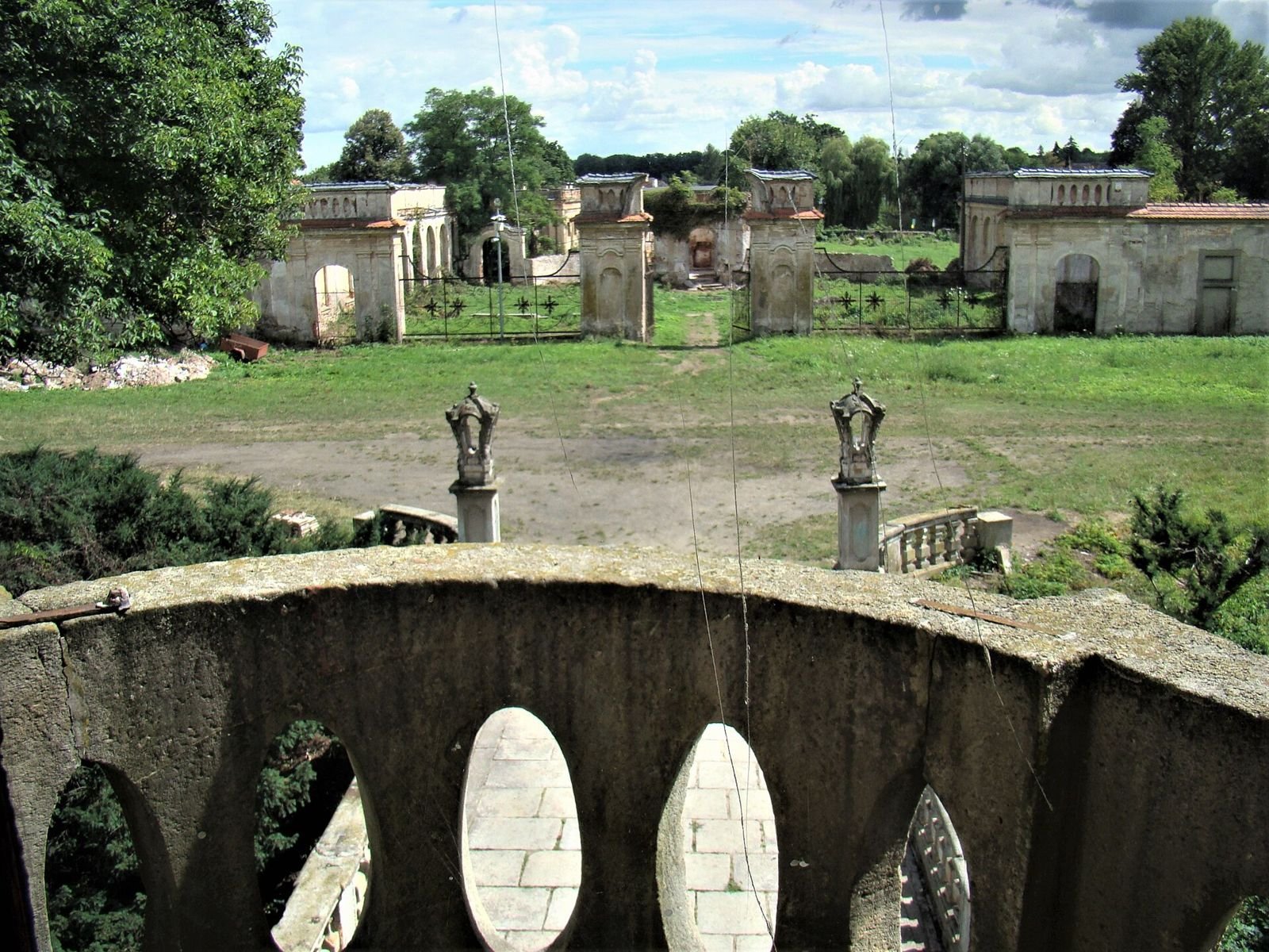 Widok z pałacu przez kamienną balustradę na bramę i zabudowania oranżerii na końcu trawiastego dziedzińca.