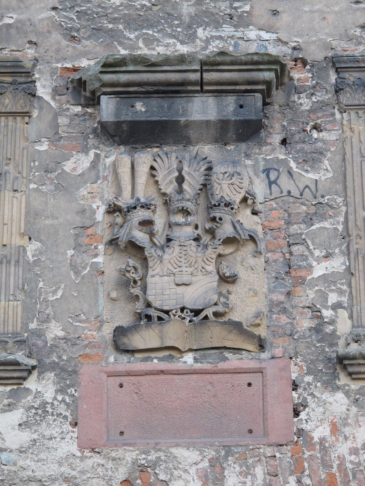 Kamienny relief herbowy w murze zamku, z tarczą herbową i prostokątną płytą pod spodem.
