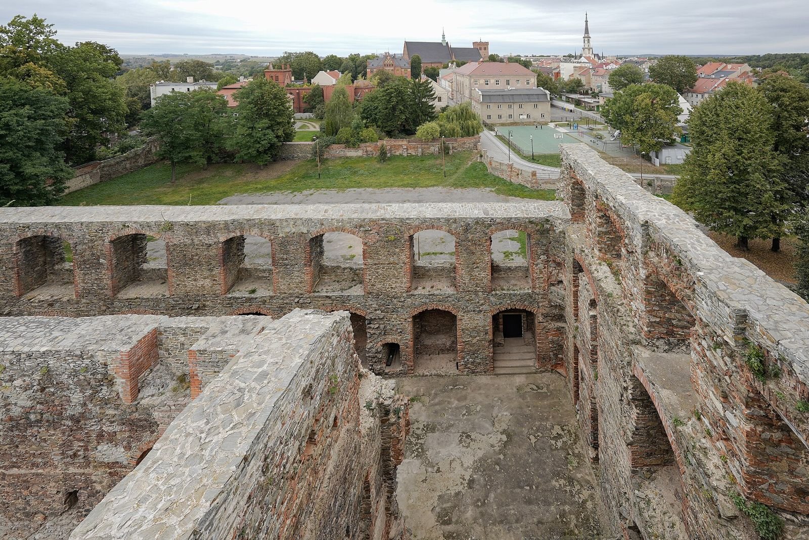 Widok z murów zamku na dziedziniec z arkadowymi wnękami i na miasto z wieżą ratusza w oddali.